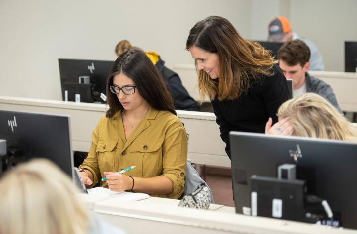 A student sits in front of a computer monitor as a teacher looks over their shoulder.